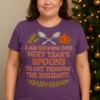Shown on Curvy Fit (XL–2XL) body wearing heather purple T-shirt with 'I am dipping into next year's spoons to get through the holidays' unseen illness awareness design, standing in a cozy Christmas living room with holiday lights