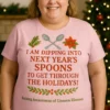 Shown on Plus Fit (3XL–5XL) body wearing light pink T-shirt with 'I am dipping into next year's spoons to get through the holidays' unseen illness awareness design, standing in a cozy Christmas living room with holiday lights and wreath