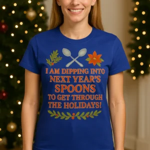Shown on Straight Fit (S–L) body wearing royal blue T-shirt with 'I am dipping into next year's spoons to get through the holidays' unseen illness awareness design, standing in a cozy Christmas living room with holiday lights and wreath