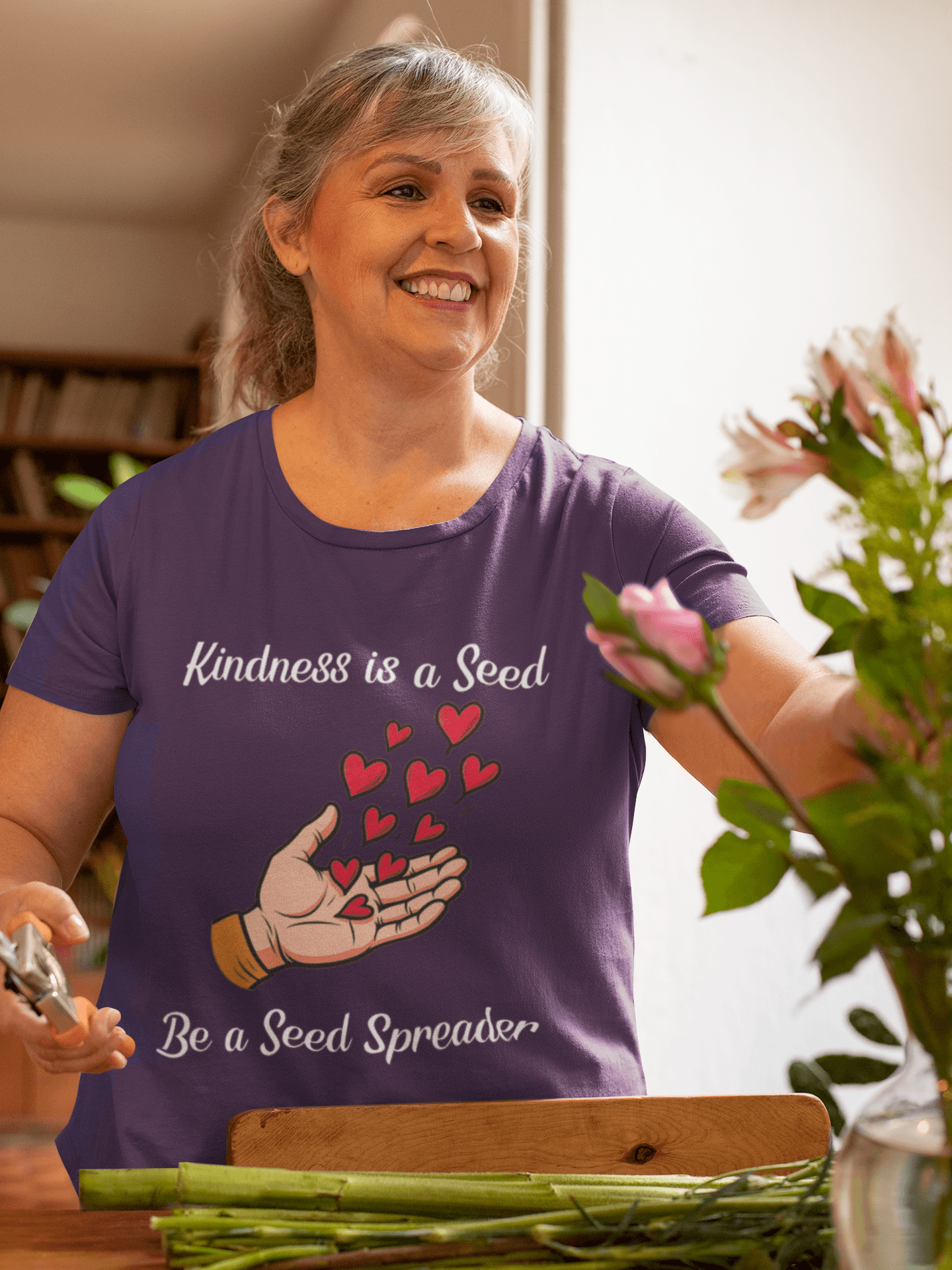 Kindness is a Seed - Be a Seed Spreader in Heather Purple Short Sleeve T-Shirt. Female model is trimming flower stems for bouquet in vase.