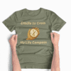 Cradle to Cross - My Life Compass in Military Green, laid out flat with hands smoothing out wrinkles