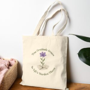 True Gratitude Grows in Life’s Hardest Places Canvas Tote in Natural on a table leaning on a wall